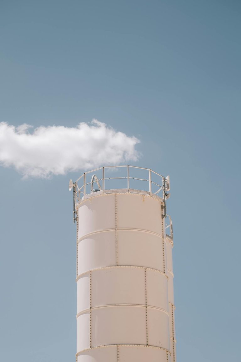 A white industrial smokestack releasing smoke into a clear blue sky, symbolizing pollution and energy production.